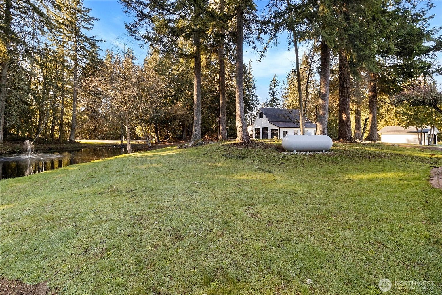 This exterior shot showcases a well-maintained yard with lush green grass, mature trees, and a tranquil pond with a fountain. A white house with a dark roof is visible in the background, along with a large propane tank. The scene evokes a sense of peaceful, private living in a natural setting.