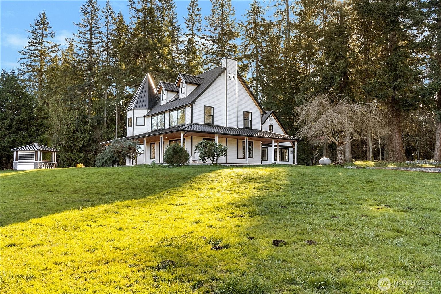 This is a front exterior view of a large, modern farmhouse-style home. The house features white siding, a black metal roof, and a prominent turret. A wide, covered porch wraps around the front and side of the house, and the home is situated on a well-manicured lawn with mature trees in the background.