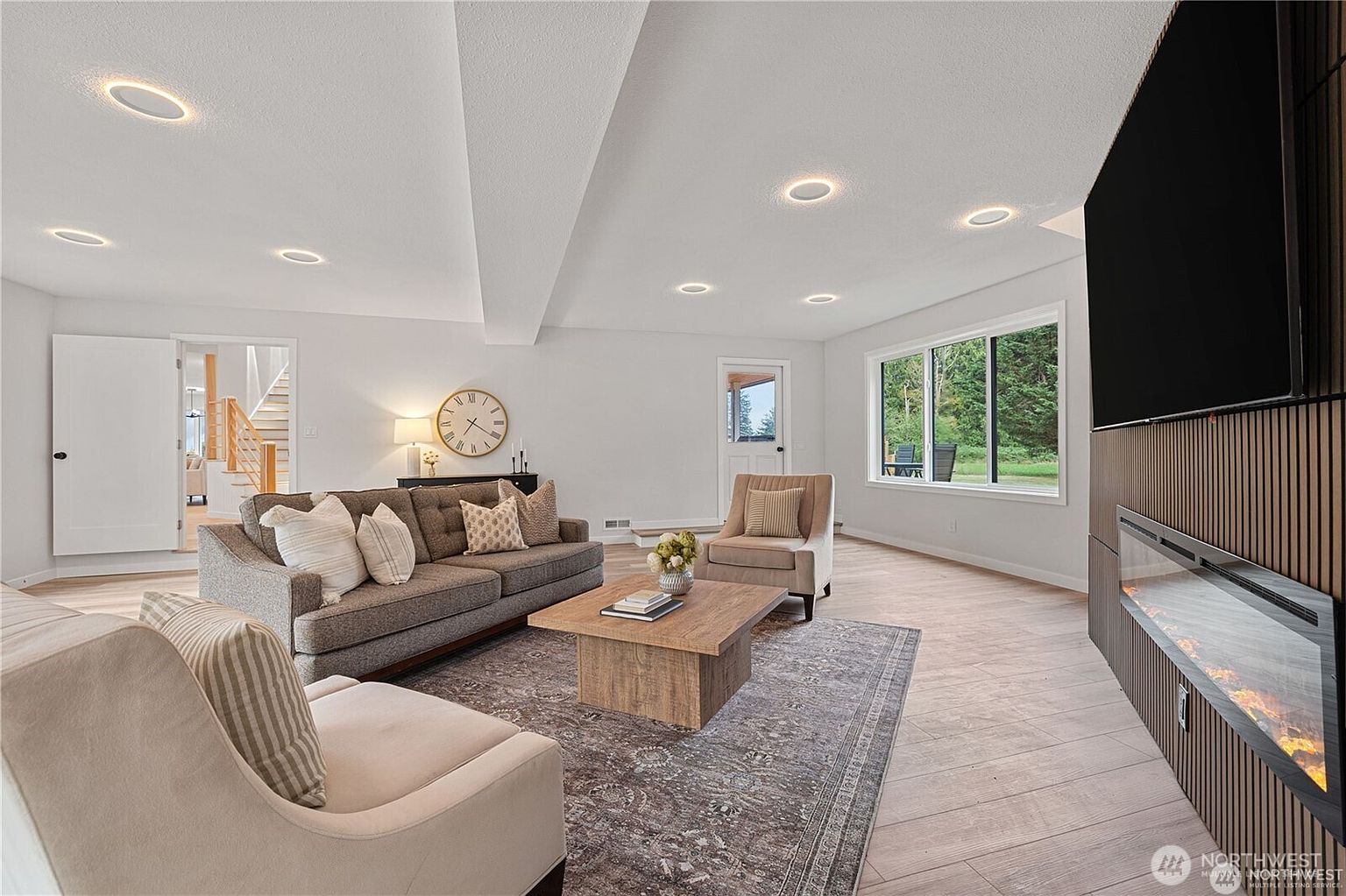 This is a well-lit living room featuring a gray sofa with decorative pillows, a light-colored armchair, and a wooden coffee table on a patterned rug. A modern fireplace and TV are mounted on the wall, and large windows provide natural light. The room has a cozy and inviting atmosphere, perfect for relaxation and entertainment.