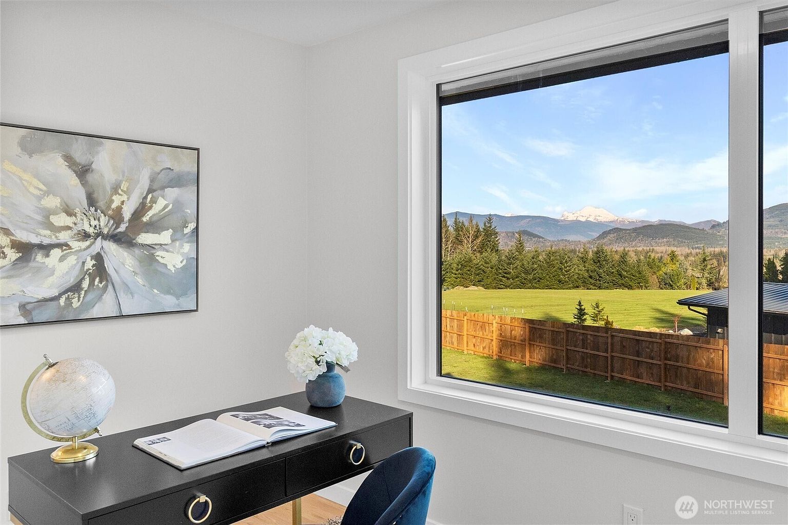 This is an interior shot of a home office featuring a dark-colored desk with gold accents, a blue velvet chair, and a globe. A large window provides a scenic view of a green field, trees, and mountains in the distance, while a framed floral painting hangs on the wall. The room has a bright and airy feel, creating a pleasant workspace.
