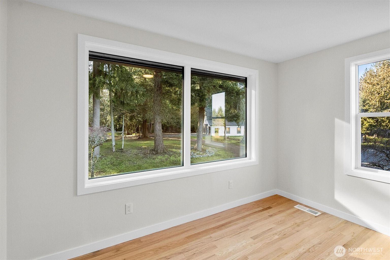 This is a bright, empty bedroom with hardwood floors and white walls, creating a clean and modern aesthetic. A large window offers a view of the trees outside, bringing natural light into the space. The room is simple and ready for personalization, making it an inviting space for potential buyers.