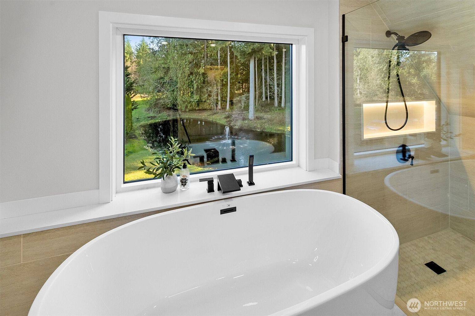 This is a luxurious primary bathroom featuring a freestanding white bathtub positioned in front of a large window with a view of a pond and trees. The bathroom also includes a glass-enclosed shower with a built-in niche. The overall impression is one of tranquility and spa-like elegance.