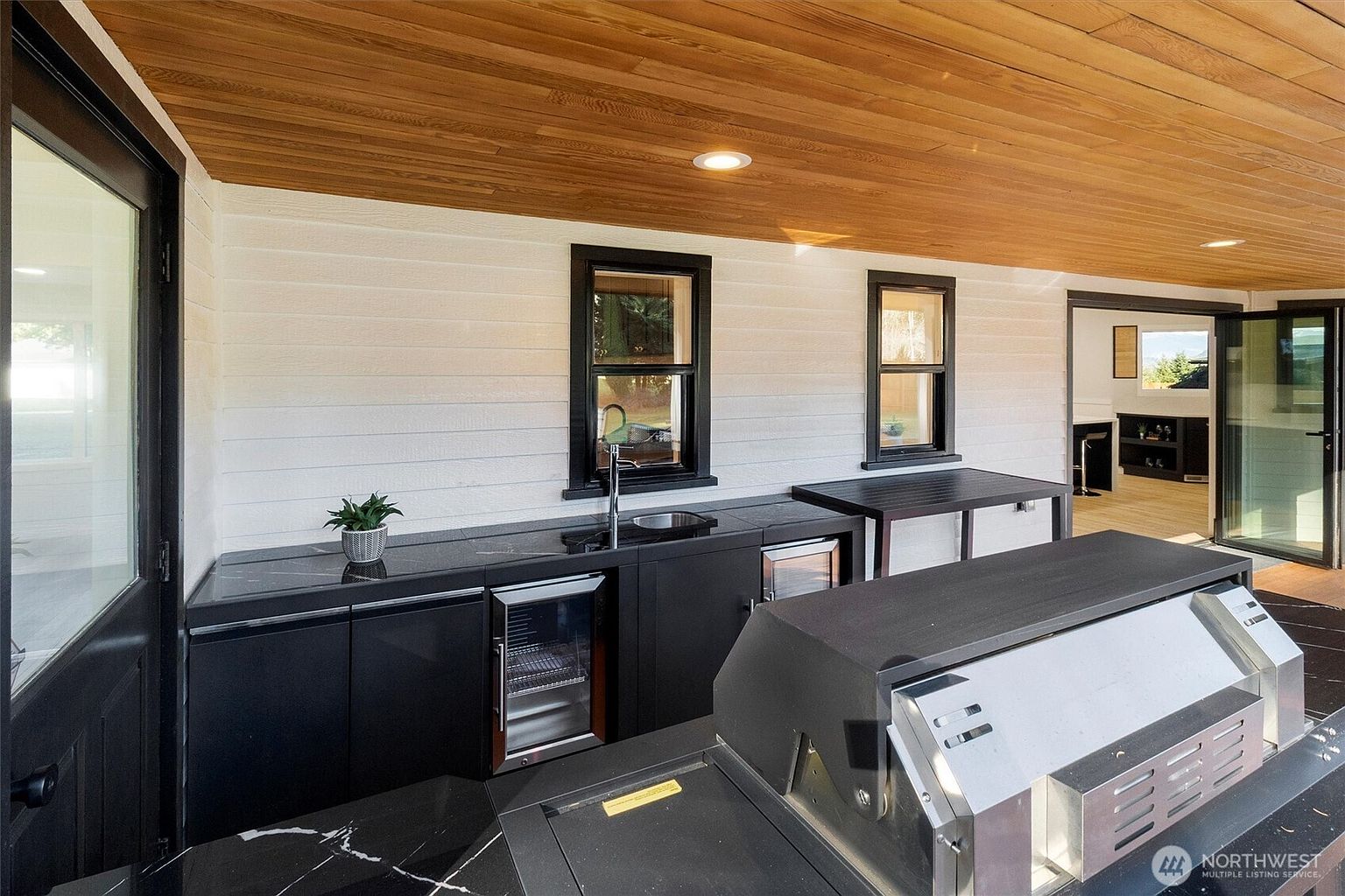 This image showcases an outdoor kitchen area on a patio or deck. The space features black cabinetry with a built-in sink, beverage cooler, and a grill. The ceiling is wood paneled, and the area is well-lit, creating an inviting and functional outdoor living space.
