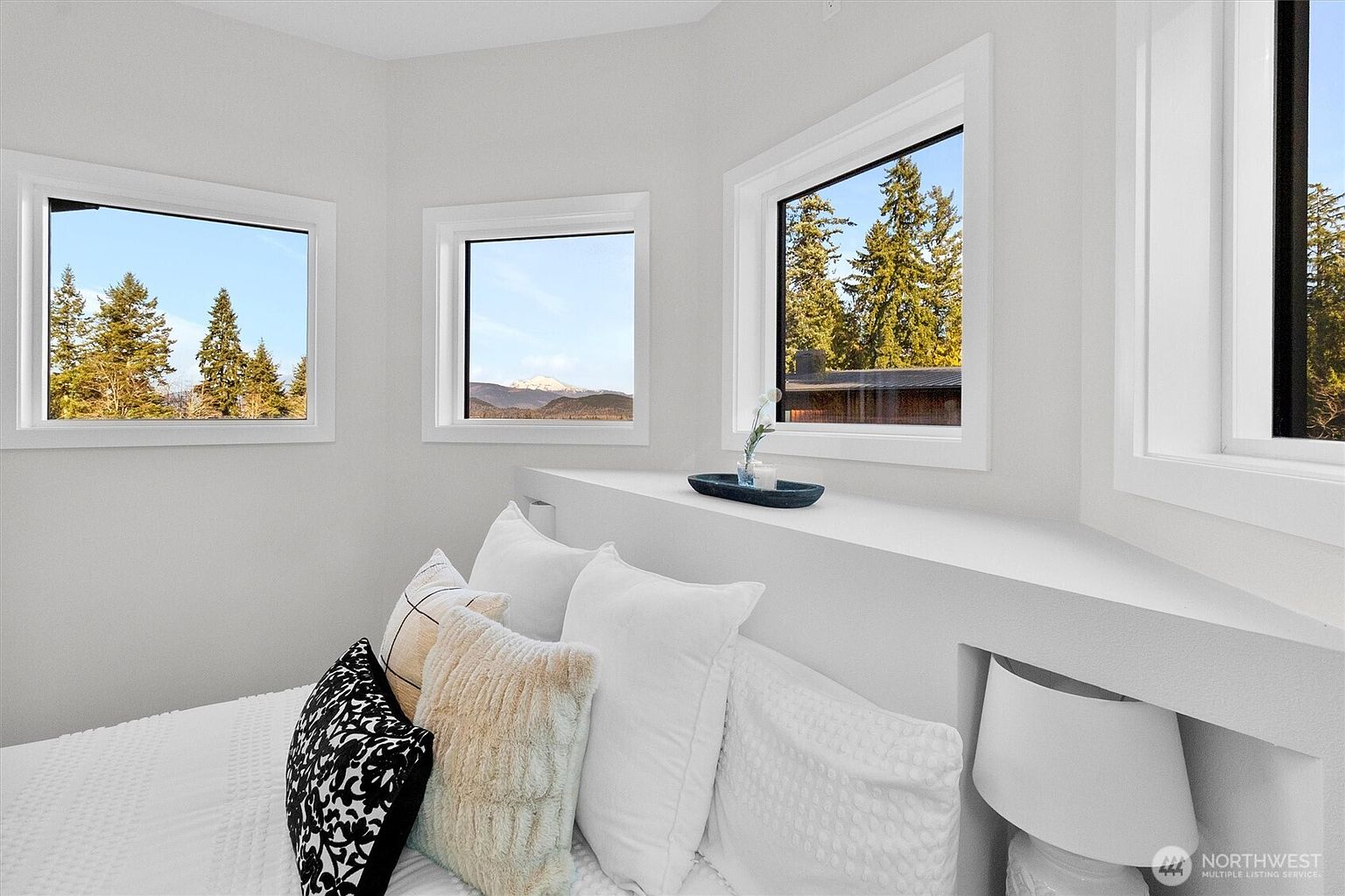 This is a bright and airy bedroom featuring a minimalist design with white walls and bedding. Three windows offer views of the surrounding landscape, bringing natural light into the space. A built-in shelf behind the bed serves as a headboard and display area, adding a touch of modern elegance.