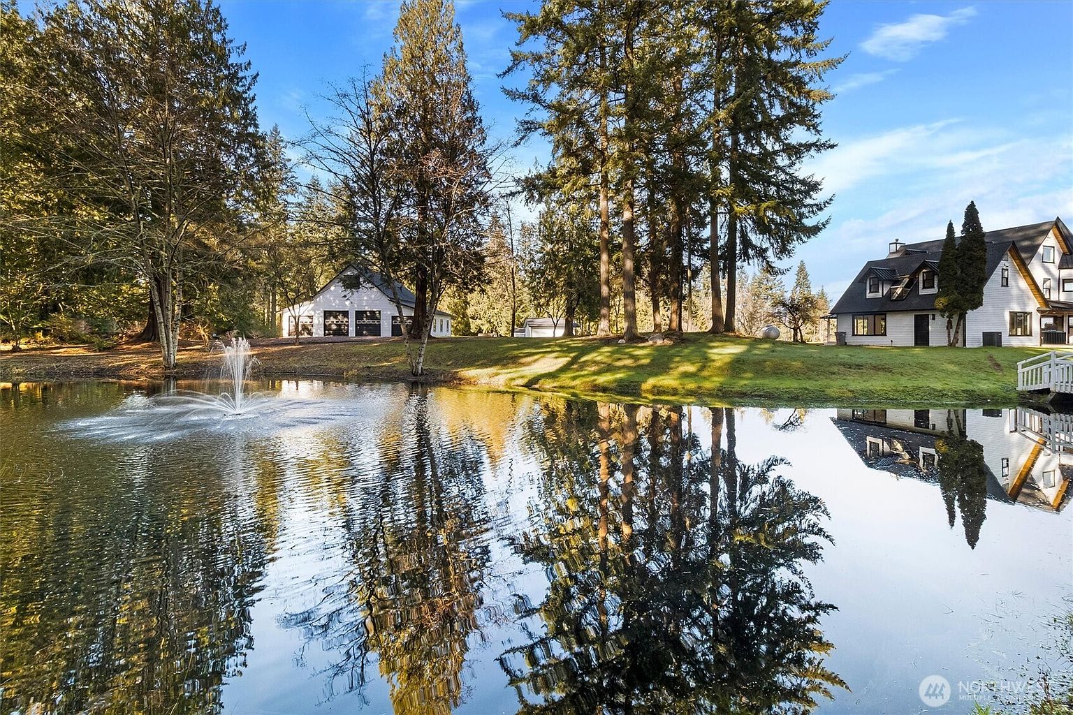 This picturesque exterior shot showcases a serene pond with a fountain, reflecting the surrounding trees and a charming white house with black accents. A well-manicured lawn leads up to the house, creating a tranquil and inviting atmosphere. The image highlights the property's natural beauty and peaceful setting.