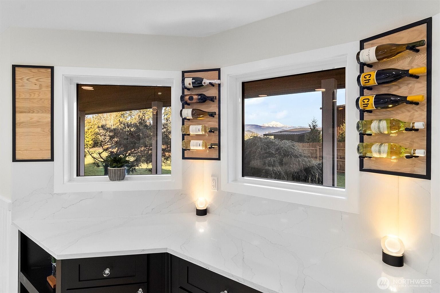 This is a bright kitchen featuring white marble countertops and black cabinetry. Two windows offer views of the outdoors, and wine racks are mounted on the wall next to each window. Small, modern lamps provide accent lighting on the countertop.