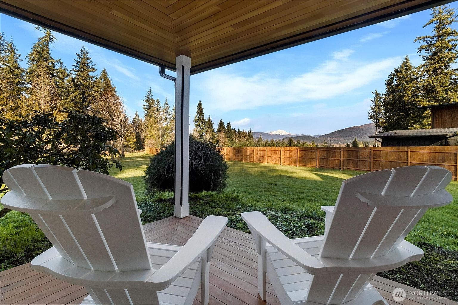 This image showcases a serene outdoor patio or deck area, featuring two white Adirondack chairs positioned to overlook a well-maintained lawn and a wooden fence. The view extends to distant mountains under a clear blue sky, creating a peaceful and inviting atmosphere. The wooden ceiling above the deck adds a touch of rustic charm to the scene.