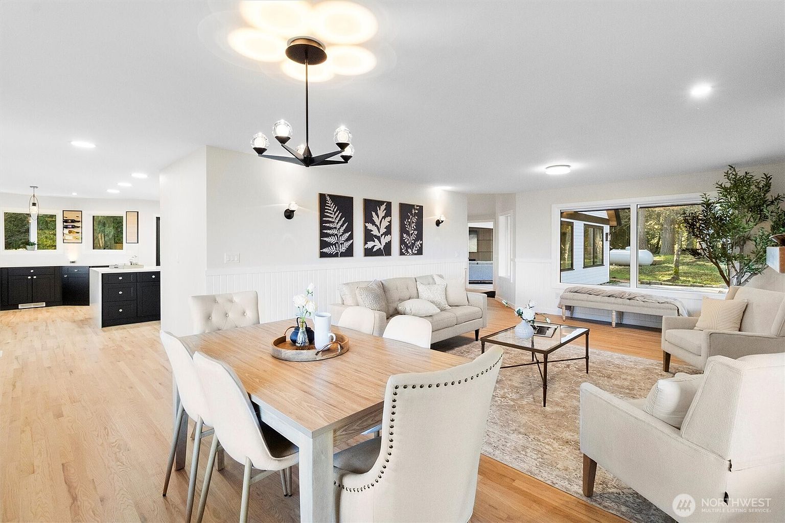 This interior shot showcases a combined dining and living area, featuring a light wood dining table surrounded by upholstered chairs, complemented by a modern chandelier. The living area includes a neutral-toned sofa, armchairs, and a coffee table, all set on a patterned rug. Large windows provide ample natural light, enhancing the open and airy feel of the space.