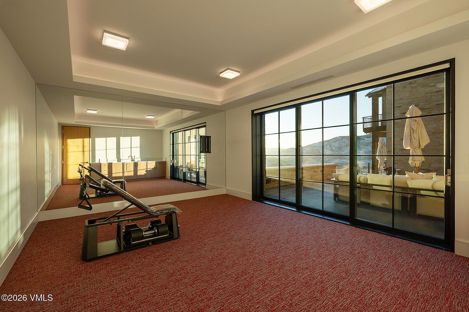 This home gym features a large mirrored wall that enhances the sense of space and a piece of exercise equipment positioned in the center. A wall of floor-to-ceiling sliding glass doors provides abundant natural light and a seamless transition to an outdoor patio with mountain views. The room is finished with red carpeting and recessed lighting, creating a functional and bright workout environment.