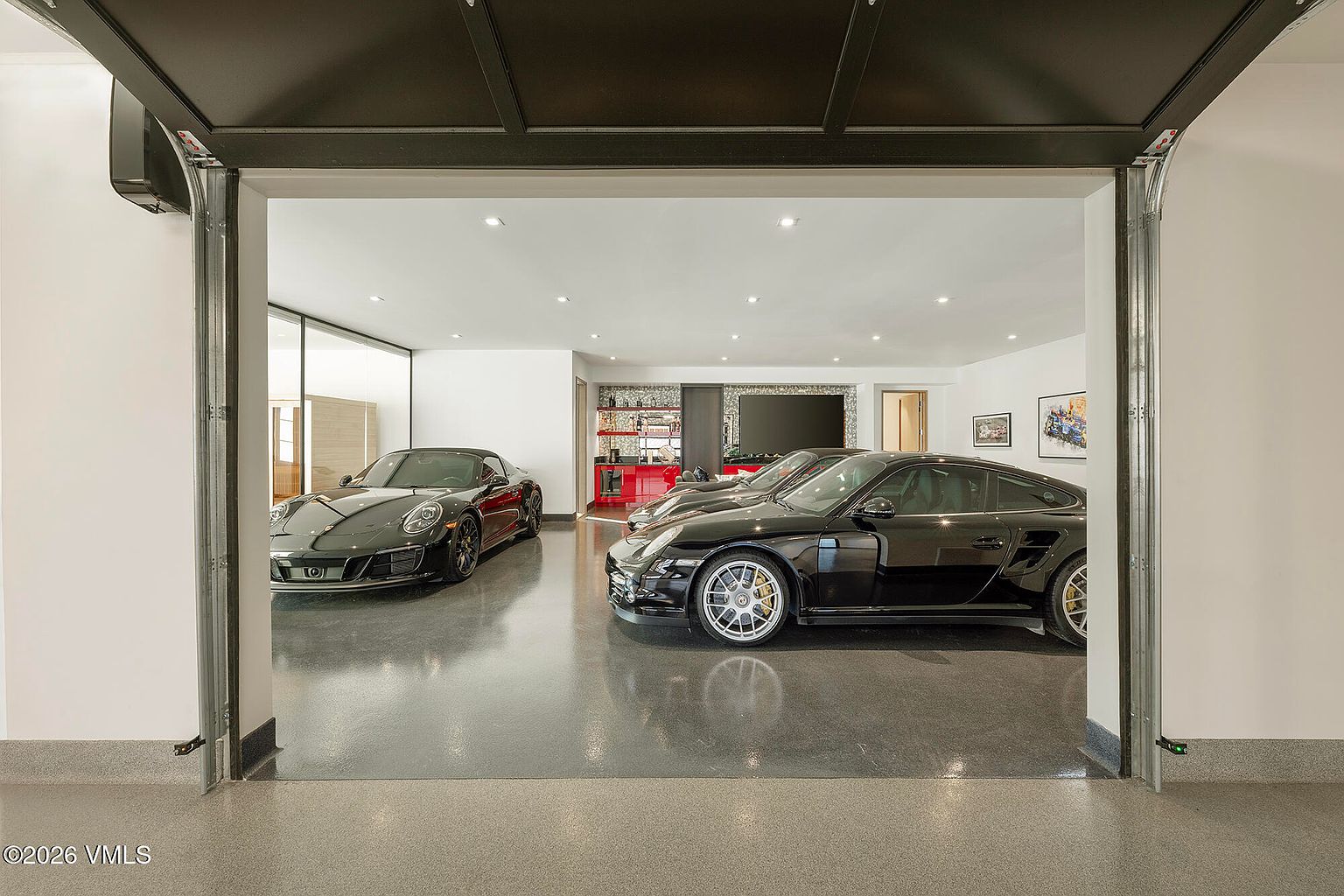 This high-end, climate-controlled garage features a polished, reflective floor and recessed lighting, creating a showroom-like atmosphere for the two luxury sports cars parked inside. The space includes a built-in bar area with a red base and glass shelving, as well as a wall-mounted television, emphasizing a lifestyle-focused design. The perspective is from the threshold of the open garage door, looking into the pristine, well-lit interior.