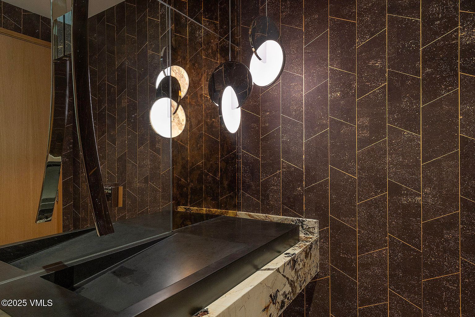 This sophisticated powder room features a striking wall treatment with dark, textured tiles arranged in a geometric herringbone pattern, accented by thin metallic lines. A sleek, dark stone vanity with a dramatic, light-veined stone edge provides a bold contrast, while three modern, circular pendant lights hang above to create a warm, ambient glow. The perspective is a close-up, eye-level shot that emphasizes the high-end materials and moody, contemporary aesthetic of the space.