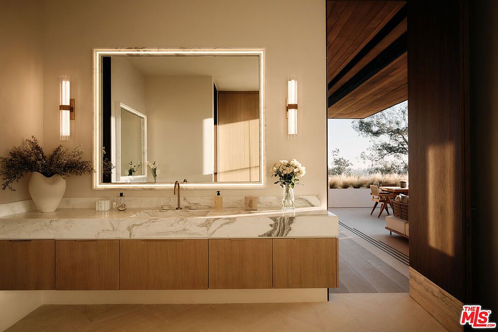 This is a luxurious primary bathroom featuring a marble countertop vanity with wood cabinets and a large, illuminated mirror. The space is well-lit with modern sconces and natural light streaming in from an adjacent outdoor patio area. The design emphasizes clean lines and high-end finishes, creating a spa-like atmosphere.