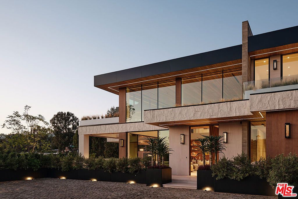 This is a front exterior view of a modern, two-story home. The house features a flat roof with a dark trim, large windows, and a combination of stone and wood siding. The landscaping includes manicured bushes in black planters and a paved driveway, creating a sophisticated and inviting curb appeal.