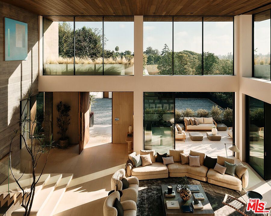 This is a spacious, modern living room with floor-to-ceiling windows offering views of the outdoor patio and greenery. The room features a large sectional sofa, a coffee table with decorative items, and a staircase with glass railings. The open floor plan and natural light create an inviting and luxurious atmosphere.