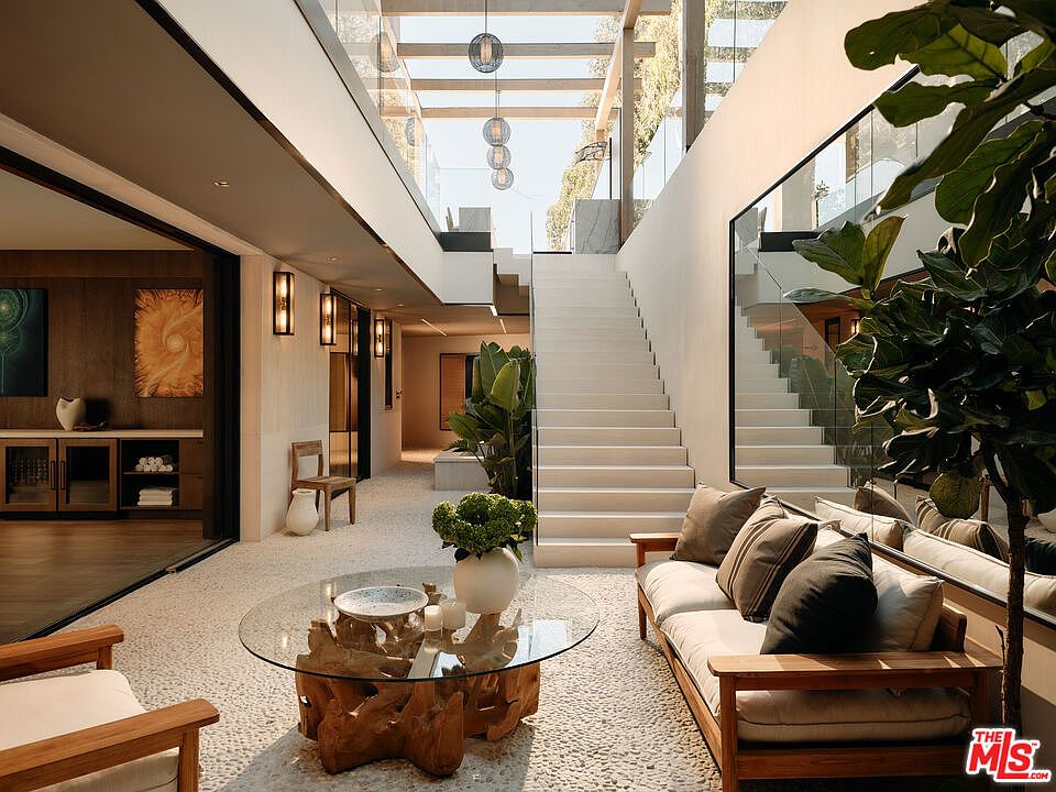 This interior shot showcases a modern hallway and staircase. The space features a unique pebble floor, a glass-topped coffee table with a driftwood base, and a comfortable-looking sofa with throw pillows. A grand staircase with glass railings leads to the upper level, and natural light floods the area through a skylight, creating a bright and airy atmosphere.