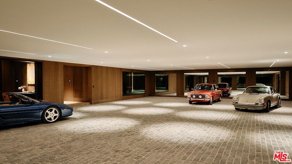 This is a spacious garage featuring a collection of classic cars, including a blue convertible, a red sedan, and a silver sports car. The garage has a modern design with wood-paneled walls, large windows, and a light-colored brick floor. The lighting is provided by recessed ceiling lights and linear fixtures, creating a bright and inviting space.