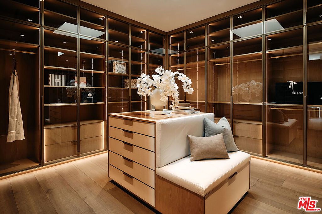 This is a luxurious walk-in closet featuring custom cabinetry with glass doors and integrated lighting. A central island with drawers and a cushioned seating area provides a space for organizing and relaxing. The hardwood flooring and neutral color palette create a sophisticated and inviting atmosphere.
