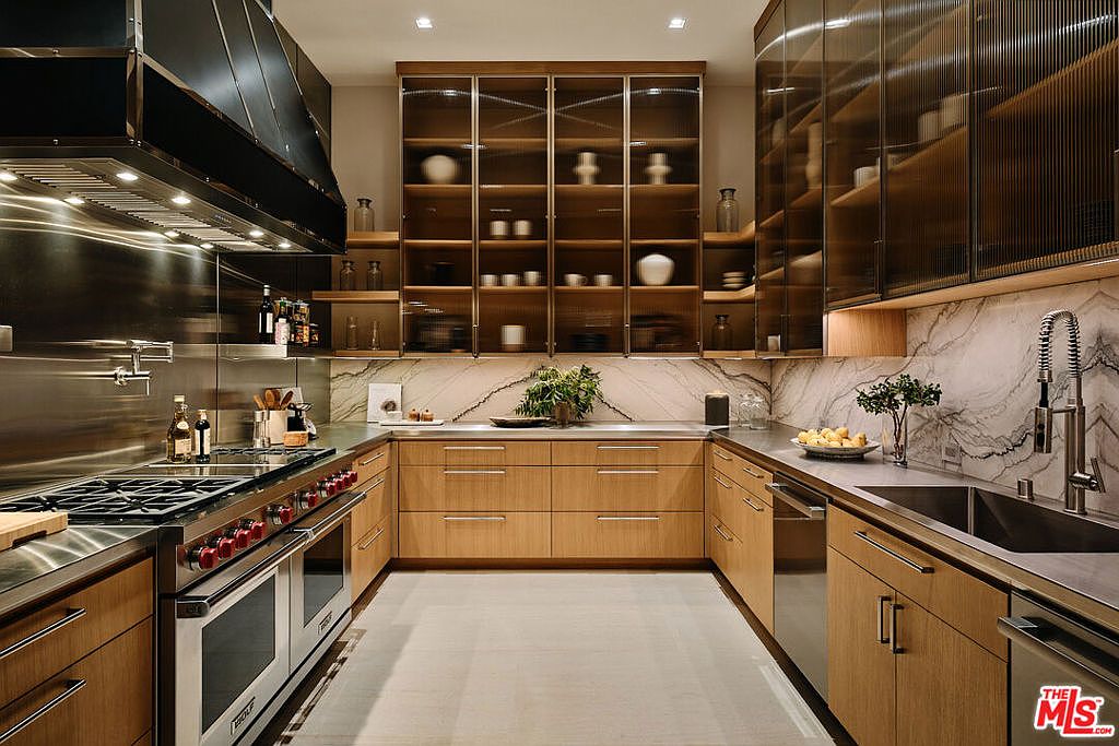 This is a well-lit, modern kitchen featuring light wood cabinetry, stainless steel appliances, and marble backsplash. The kitchen has a U-shaped layout with a large range and hood, as well as glass-front cabinets displaying decorative items. The perspective is a medium shot, showcasing the entire kitchen layout and design.