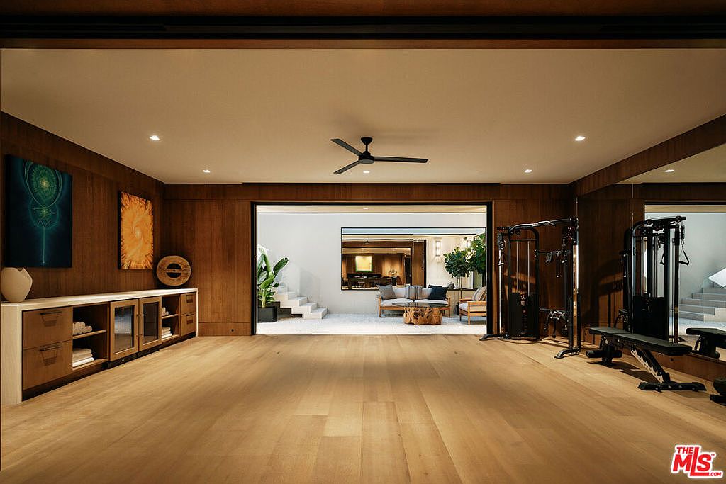 This is a spacious home gym featuring wood flooring, wood paneled walls, and a ceiling fan. The room includes a weight training machine, a bench, and a built-in cabinet with display shelves. An open doorway leads to an outdoor patio area with seating, creating a seamless indoor-outdoor flow.