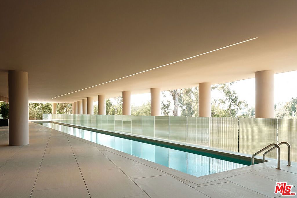 This image showcases a modern, covered pool area. The pool is long and rectangular, surrounded by a tiled deck and a low, translucent barrier. The space is supported by evenly spaced columns, creating an open yet sheltered environment, with glimpses of greenery visible beyond the structure.