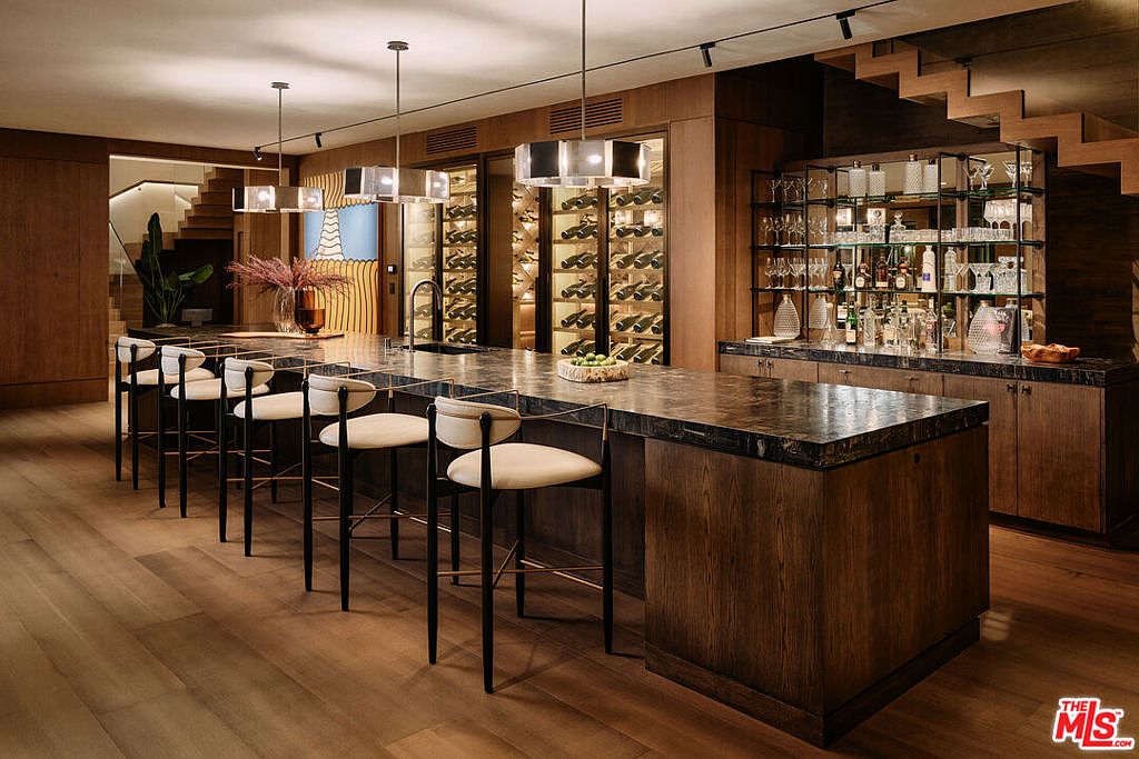 This interior shot showcases a sophisticated wine cellar and bar area. The space features a long, dark countertop with bar stools, wine storage behind glass doors, and a mirrored bar display with glassware and bottles. The warm wood tones and modern lighting create an inviting and luxurious atmosphere, perfect for entertaining.