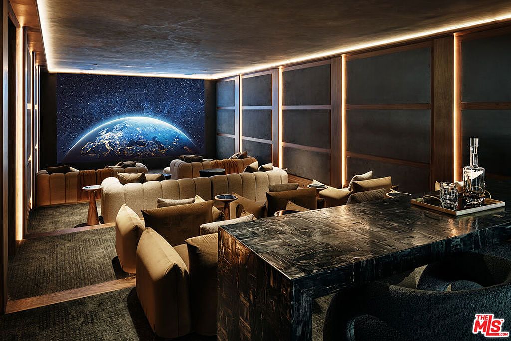 This is a luxurious home theater featuring tiered seating with plush couches and pillows, a large screen displaying a celestial image, and a dark, sophisticated color palette. The room includes a bar area with a sleek countertop and stylish chairs, creating an inviting space for entertainment. The overall impression is one of comfort and high-end design, perfect for a cinematic experience.