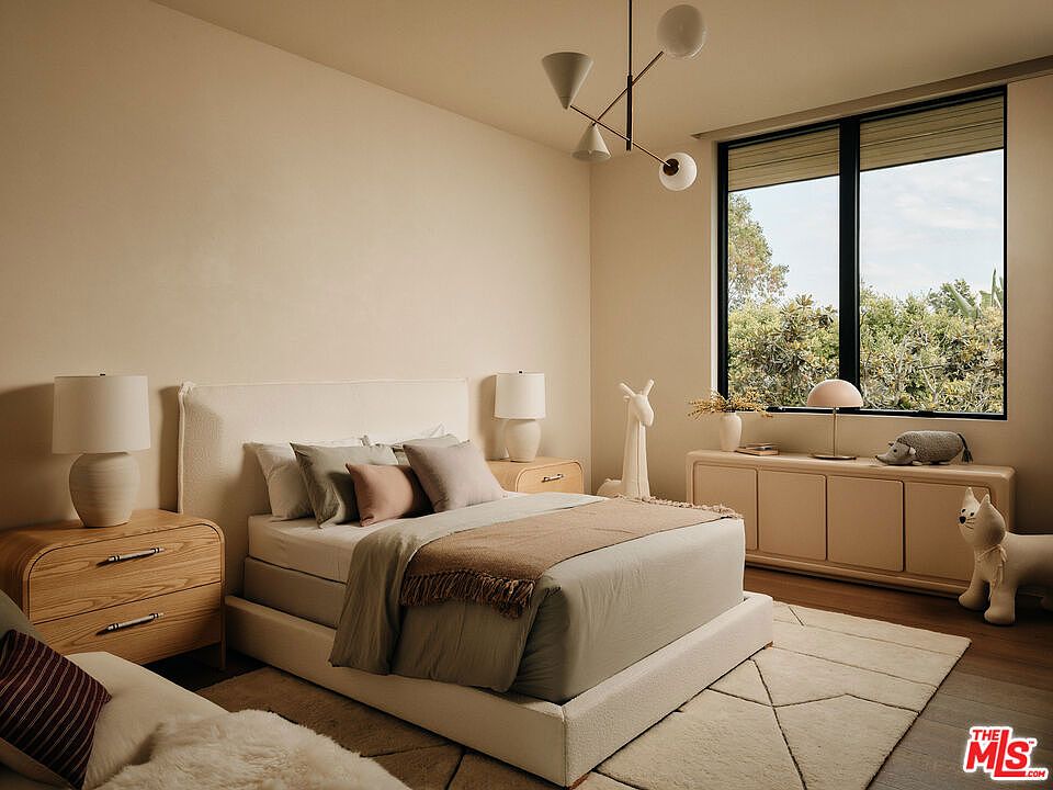 This is a serene primary bedroom featuring a neutral color palette, creating a calming and inviting atmosphere. The room includes a large bed with plush pillows and a cozy throw, bedside tables with lamps, and a dresser with decorative objects. A large window provides natural light and a view of the outdoors, enhancing the room's tranquil ambiance.