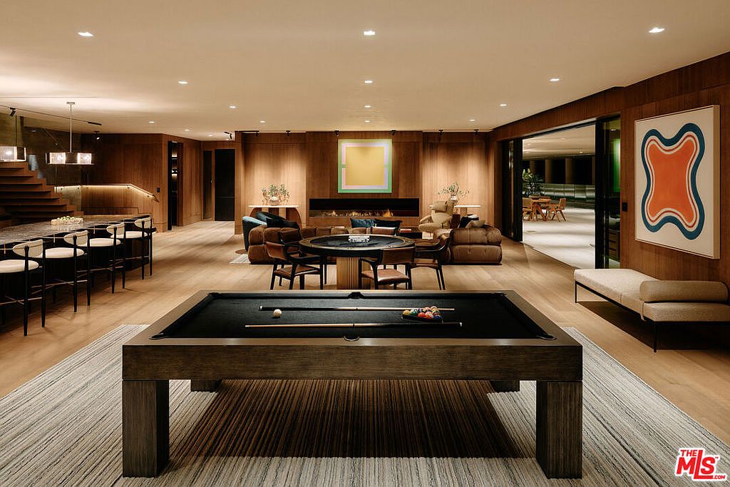 This is a spacious and luxurious living room featuring a pool table in the foreground and a seating area with a fireplace in the background. The room is decorated with modern art and has wood paneling, creating a warm and inviting atmosphere. The open floor plan leads to an outdoor area, enhancing the sense of space and connection to the outdoors.
