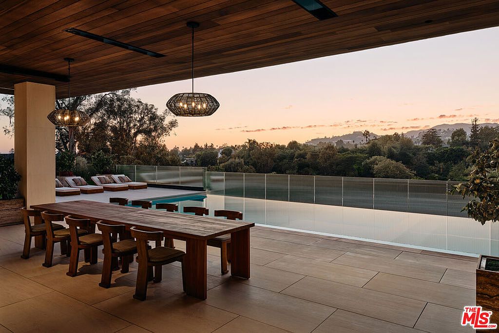 This image showcases a luxurious outdoor patio area, featuring a long wooden dining table with chairs, positioned next to a stunning infinity pool. The patio is elegantly designed with large format tiles, and the view extends to a lush landscape under a vibrant sunset sky. The scene evokes a sense of high-end living and sophisticated outdoor entertainment.