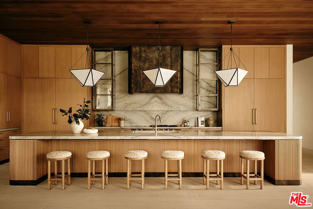 This is a well-lit, modern kitchen featuring light wood cabinetry and a large island with seating. The kitchen is equipped with stainless steel appliances and unique pendant lighting. The overall design is sleek and contemporary, creating a warm and inviting atmosphere.