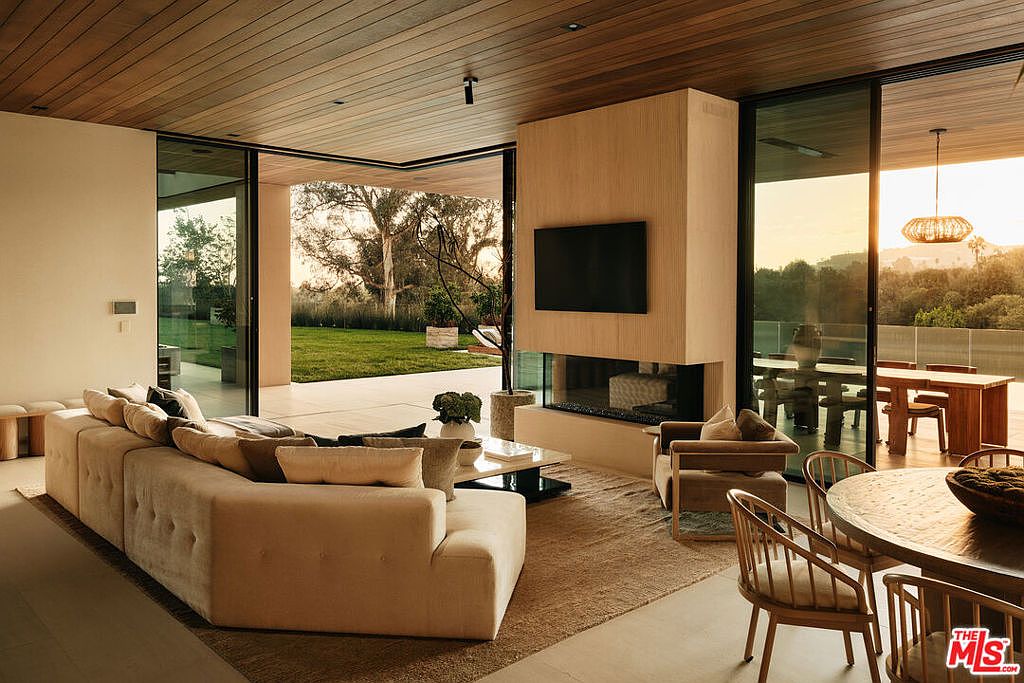 This is an interior shot of a modern living room featuring a large sectional sofa, a fireplace with a mounted television, and floor-to-ceiling glass doors that open to an outdoor area. The room has a neutral color palette with wood accents on the ceiling and fireplace surround, creating a warm and inviting atmosphere. The perspective is from a medium distance, showcasing the open floor plan and seamless transition between indoor and outdoor spaces.