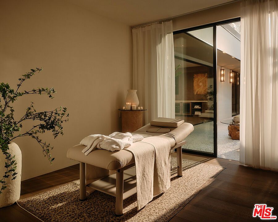 The image showcases a serene spa room featuring a massage table draped with towels, positioned on a patterned rug. Natural light filters through sheer curtains, illuminating the room and highlighting a vase with greenery. A glimpse of an outdoor area is visible through a sliding glass door, enhancing the tranquil atmosphere.