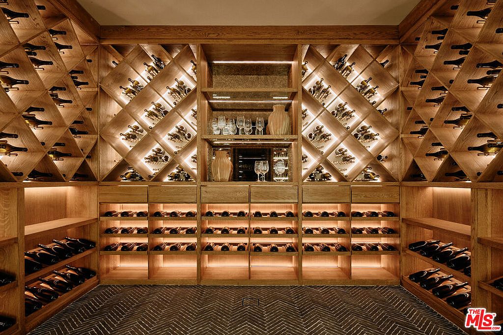 This is a well-lit wine cellar featuring custom wooden shelving designed to hold numerous wine bottles. The shelves are arranged in a diamond pattern and straight rows, with integrated lighting that highlights the bottles and glassware. The overall impression is one of sophistication and organization, perfect for a wine enthusiast.