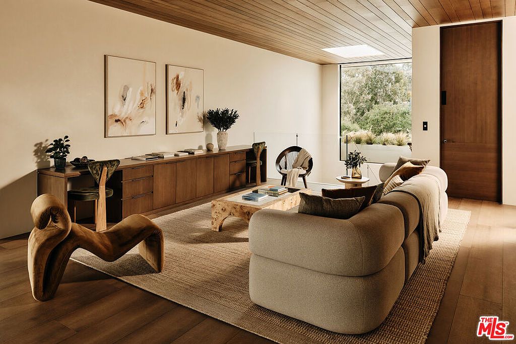 This is a stylish living room featuring a neutral color palette, wood accents, and modern furniture. The room includes a large sofa, unique sculptural chair, a wooden cabinet with artwork above, and a coffee table with books. A large window provides natural light and a view of the outdoors, enhancing the room's serene and sophisticated atmosphere.