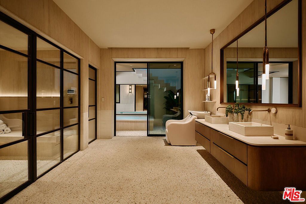 This is a luxurious primary bathroom featuring a double vanity with modern sinks and fixtures, complemented by stylish pendant lighting and a large mirror. The room includes a sauna behind a glass enclosure and a separate area with a modern chair, possibly for relaxation or grooming. The design incorporates natural tones and textures, creating a spa-like atmosphere.