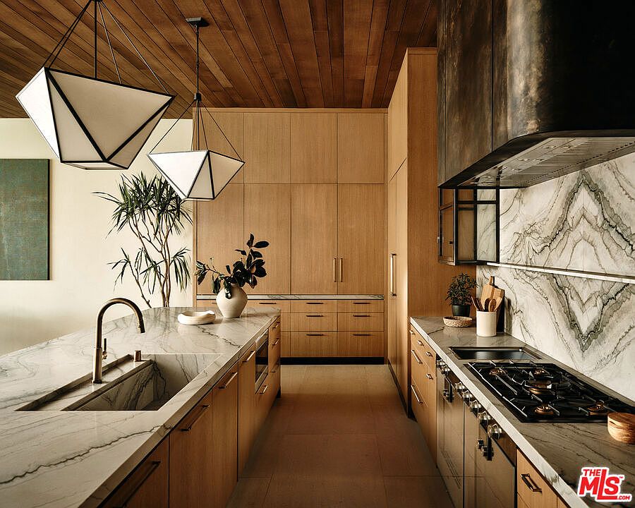 This is a well-lit, modern kitchen featuring light wood cabinetry and marble countertops. The kitchen is equipped with high-end appliances and fixtures, including a sleek cooktop and a stylish range hood. Geometric pendant lights hang above the island, adding a contemporary touch to the space.