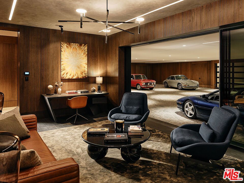 This image showcases a luxurious living room that seamlessly transitions into a garage space featuring classic cars. The living area is furnished with a leather sofa, stylish armchairs, and a modern coffee table, complemented by a sleek desk area with an abstract art piece. The design emphasizes a blend of comfort and sophistication, making it an ideal space for relaxation and showcasing a passion for automobiles.