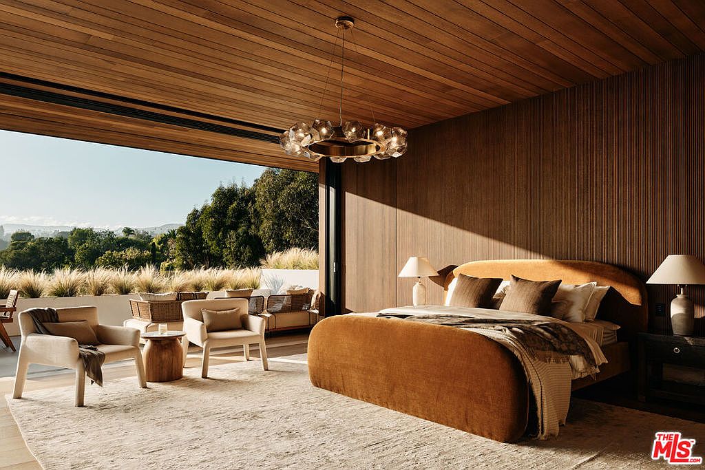 This is a luxurious primary bedroom featuring a large bed with a brown velvet headboard and matching bed frame, complemented by a modern chandelier. The room opens to an outdoor patio with seating, offering a seamless indoor-outdoor living experience. The walls and ceiling are clad in wood, creating a warm and inviting atmosphere.