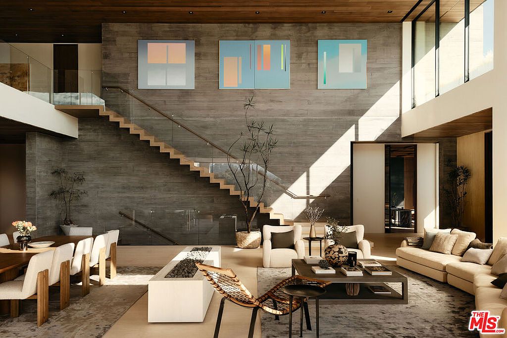 This is a spacious living room featuring a modern design with a focus on natural light and textures. The room includes a staircase with glass railings, a large concrete wall adorned with abstract art, and comfortable seating arrangements. A fireplace and various plants add to the ambiance, creating a sophisticated and inviting space.
