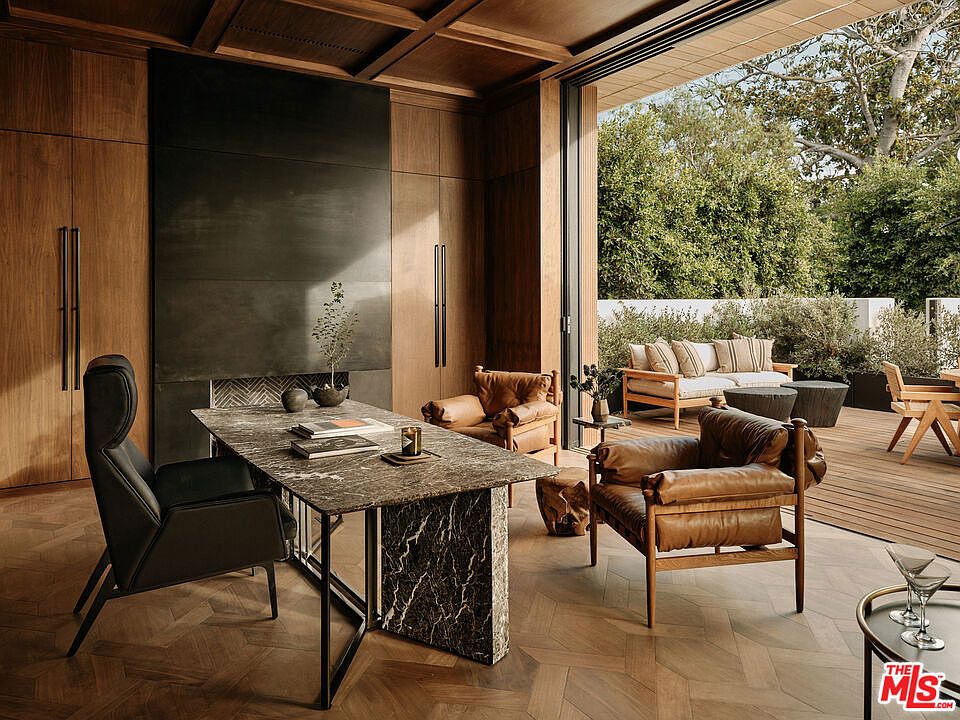 This image showcases a sophisticated home office with a seamless indoor-outdoor transition. The interior features rich wood paneling, a striking dark stone desk, and comfortable leather seating, while the open doorway leads to a furnished patio with lush greenery. The design emphasizes natural light and a connection to nature, creating a serene and productive workspace.
