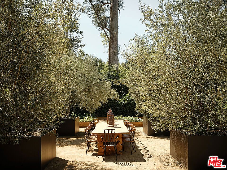 This image showcases a beautifully designed outdoor dining area, perfect for entertaining. A long wooden table is surrounded by chairs, creating an inviting space for gatherings. The area is framed by large potted trees, adding a sense of privacy and elegance to the outdoor setting.