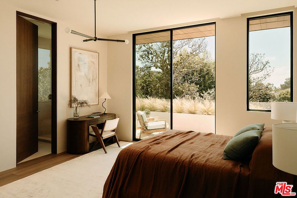 This is a bright and airy primary bedroom featuring a large bed with a brown duvet and green pillows, a modern desk with a white chair, and large windows offering views of the outdoor greenery. The room is decorated in neutral tones, creating a serene and inviting atmosphere. The perspective is from the foot of the bed, showcasing the room's layout and connection to the outdoors.