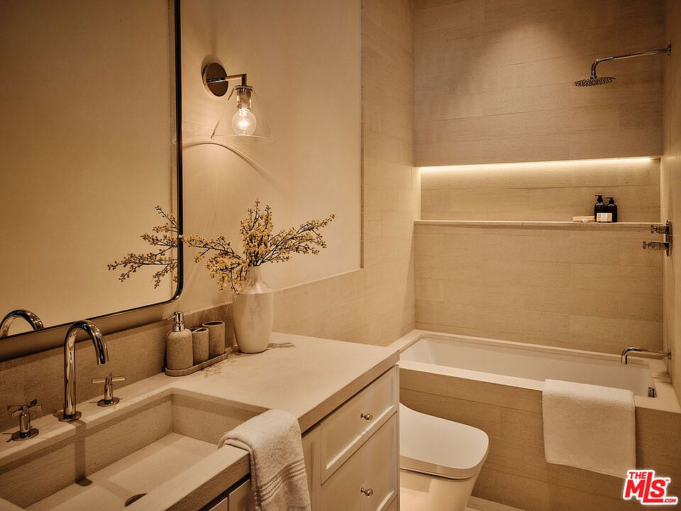 This is a well-lit primary bathroom featuring a modern design with neutral tones. The bathroom includes a large mirror, a vessel sink with chrome fixtures, and a bathtub with a shower. The walls are tiled, and there is a built-in shelf with accent lighting above the tub, creating a spa-like atmosphere.