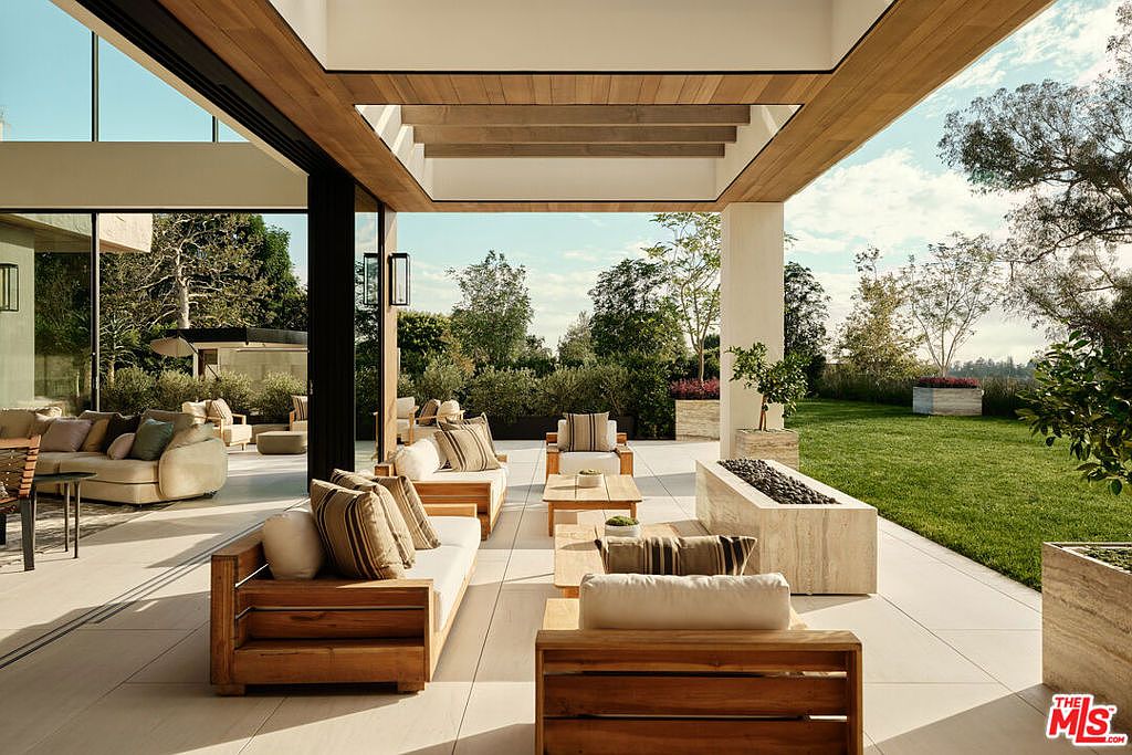This image showcases a luxurious outdoor patio area, featuring modern wooden furniture with plush cushions arranged for comfortable seating. The patio seamlessly extends into a well-manicured lawn and garden, creating a serene and inviting outdoor living space. A contemporary fire pit adds a touch of elegance and warmth to the setting.