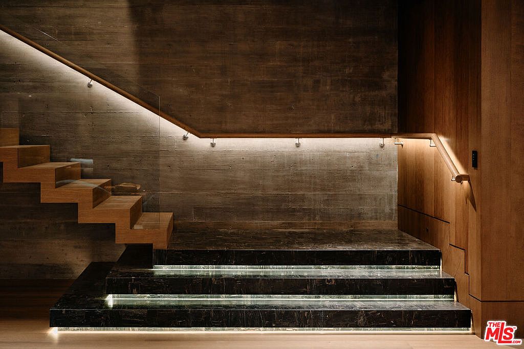This interior shot showcases a modern staircase with floating wooden steps and integrated lighting. The staircase features a glass railing and dark marble base steps with underlighting, set against a textured concrete wall and wood paneling. The overall design exudes a sophisticated and minimalist aesthetic.
