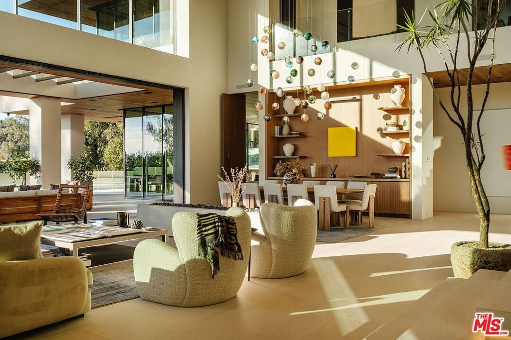 This is an interior shot of a luxurious living room featuring an open floor plan with a dining area. The room is bathed in natural light, highlighting the modern design elements such as the unique chandelier, neutral-toned furniture, and a decorative tree. Large sliding glass doors open to an outdoor space, creating a seamless indoor-outdoor living experience.