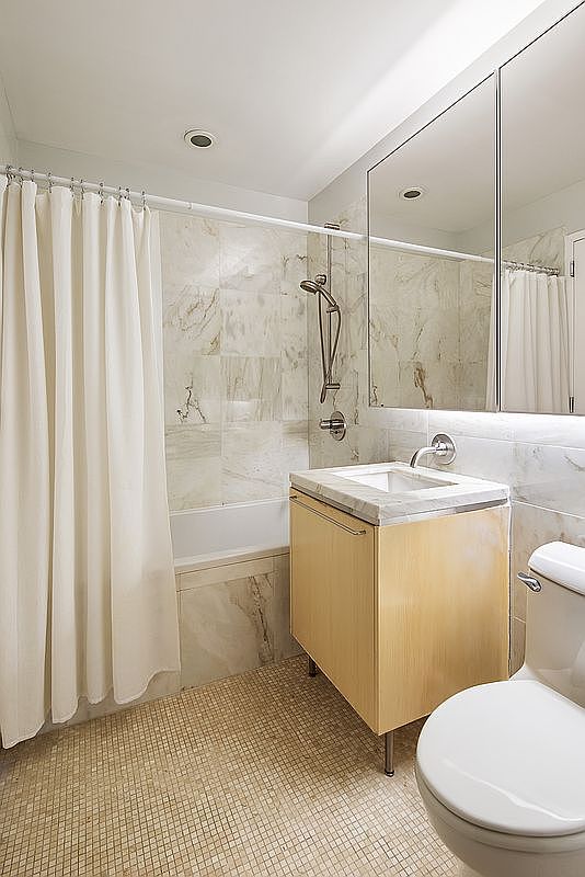 This is a well-lit bathroom featuring a shower/tub combination with a white shower curtain, a vanity with a light wood cabinet and marble countertop, and a toilet. The walls and floor are tiled in a neutral color, creating a clean and modern aesthetic. The perspective is from the doorway, showcasing the entire space.