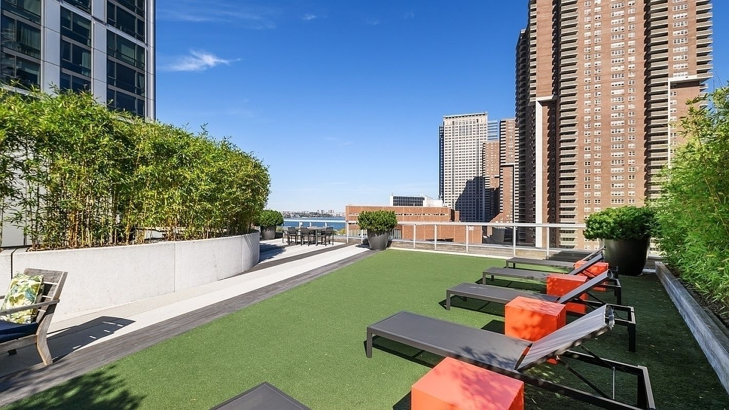 This image showcases a luxurious rooftop patio or balcony, featuring artificial turf, modern lounge chairs with vibrant orange accents, and lush greenery in planters. The space offers stunning city views, with skyscrapers visible in the background, suggesting a high-end urban living experience. The overall impression is one of sophisticated relaxation and outdoor enjoyment.