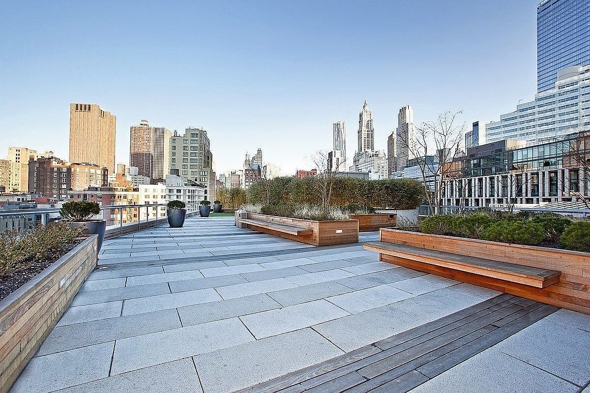This image showcases a modern rooftop patio with stunning city views. The patio features sleek gray pavers, built-in wooden planters with greenery, and integrated wooden benches for seating. The overall impression is one of urban sophistication and relaxation.