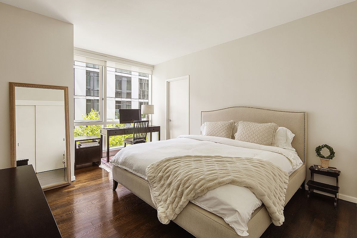 This is a well-lit primary bedroom featuring a large bed with a neutral-toned headboard and a cozy throw blanket. A desk and chair are positioned near the window, creating a functional workspace. The room is decorated in a minimalist style with hardwood floors and a full-length mirror, offering a serene and inviting atmosphere.